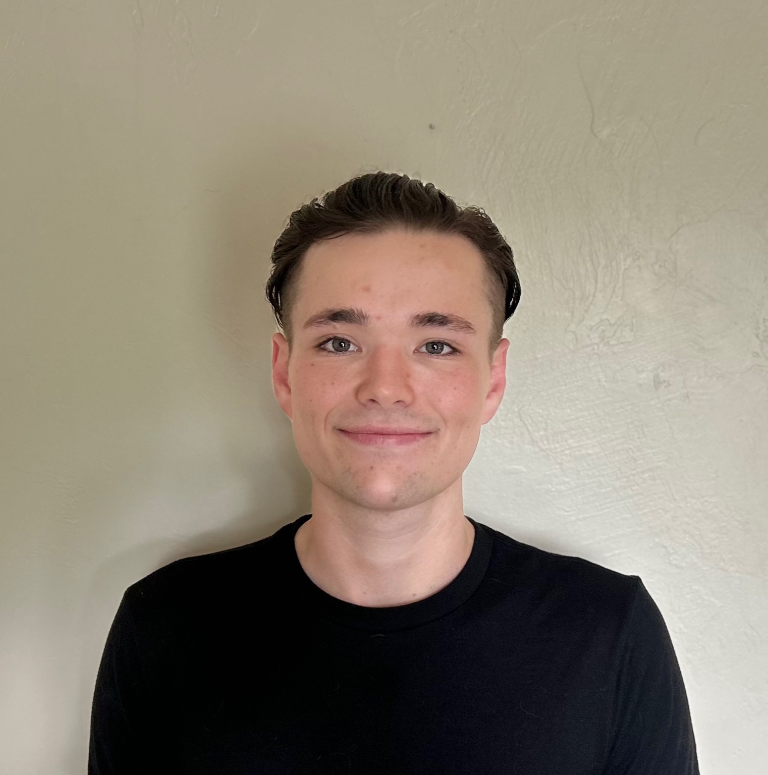 A young man in a black t-shirt stands against a plain wall, smiling slightly at the camera