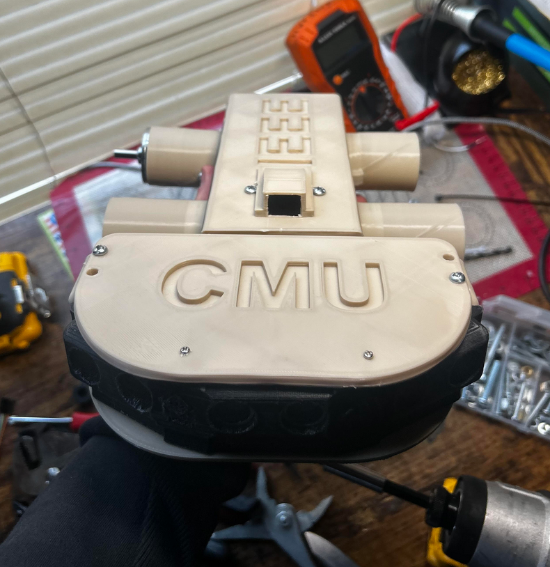 A beige 3D-printed rover chassis labeled “CMU” and “EEE,” mounted on a black base, surrounded by tools like a multimeter, screwdriver, and drill bits