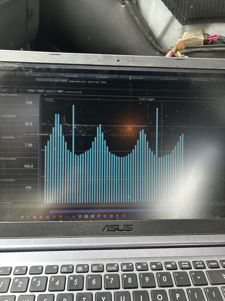 An ASUS laptop displays a blue bar graph labeled “Tooth Logger” on its screen, showing engine RPM data with visible wiring and car interior in the background