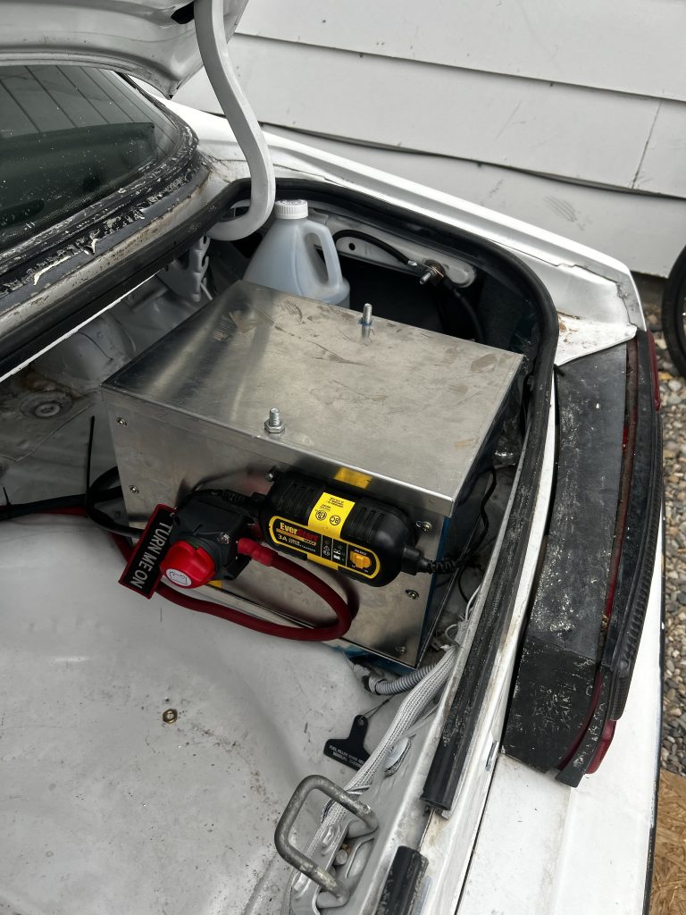 A stainless steel battery box in a car trunk, connected to a red cutoff switch and yellow-labeled EverStart battery