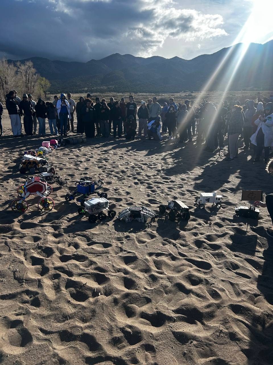 A crowd of spectators gathered behind a row of competing rovers of all shapes lined up on the dunes, with mountains and low sun in the background