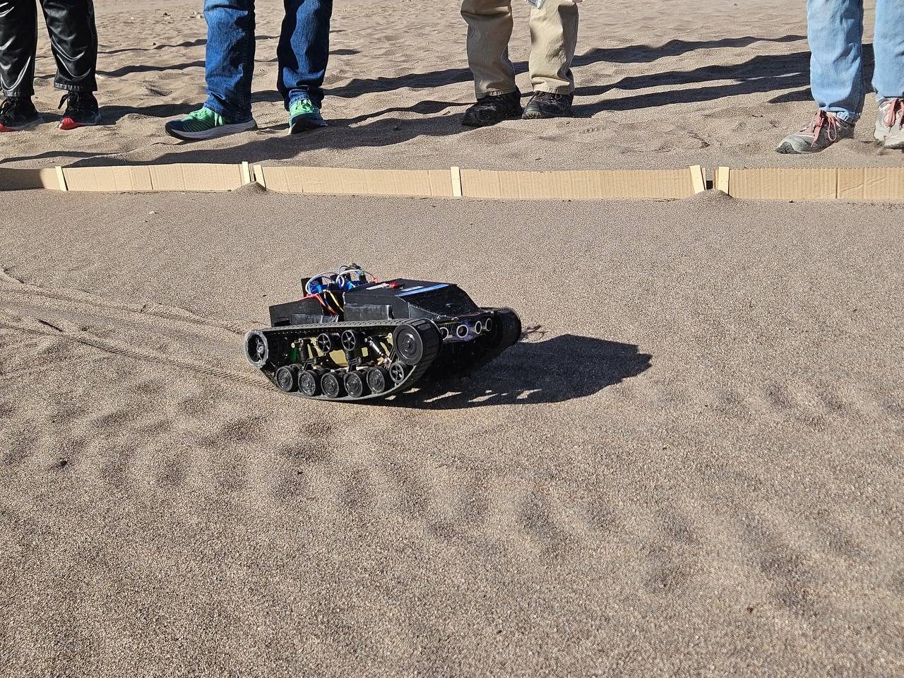 Close-up of the tracked rover with its bogie wheels and front ultrasonic sensors, crossing sand beside the cardboard lane wall