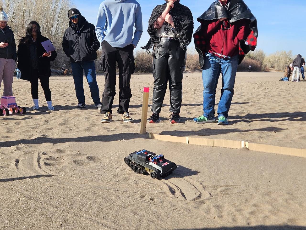 The black tracked rover driving across rippled sand inside a lane marked by a low cardboard wall, with spectators standing behind it