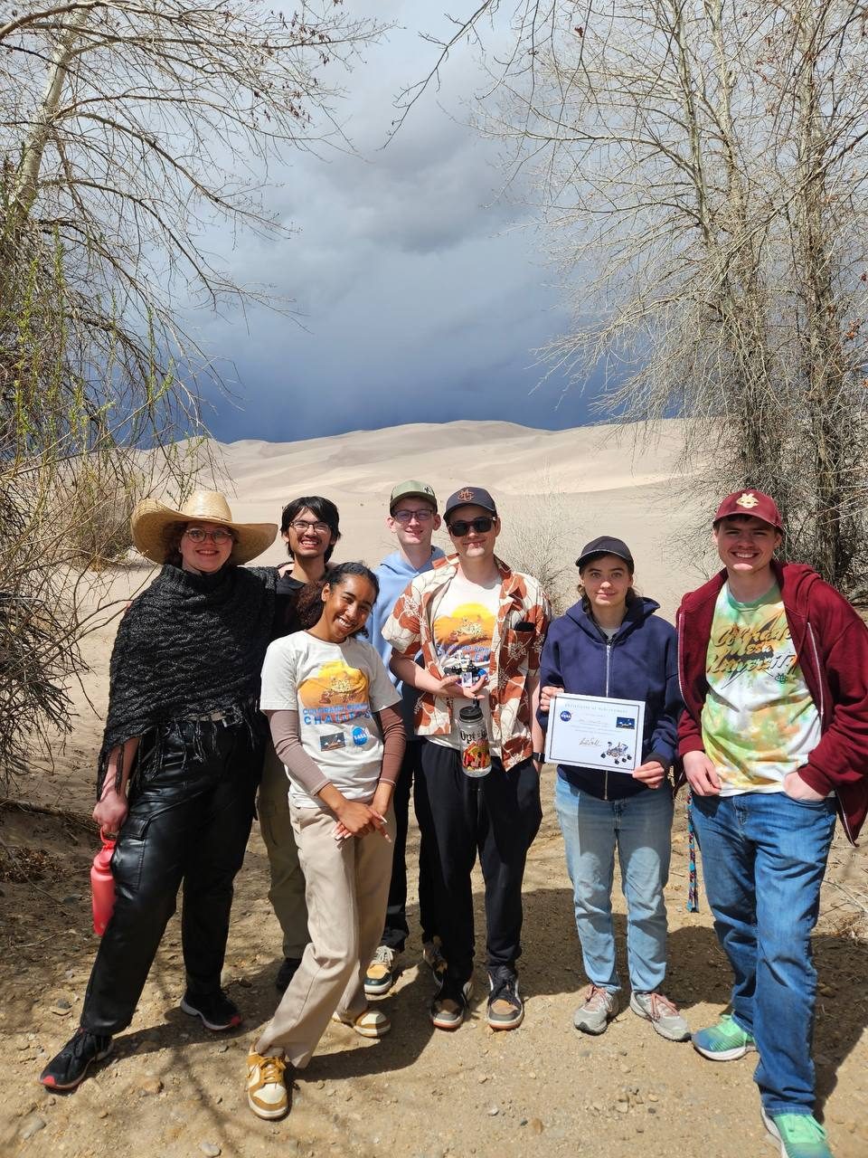 The team posing with the award certificate between bare trees, with a dark storm cloud over the dunes behind them