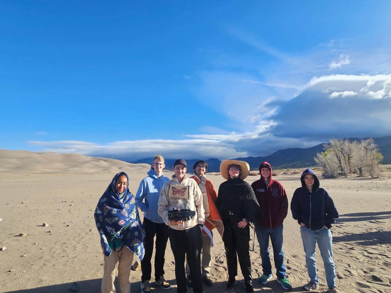Seven team members standing in front of the sand dunes, one holding the small rover, under a deep blue sky