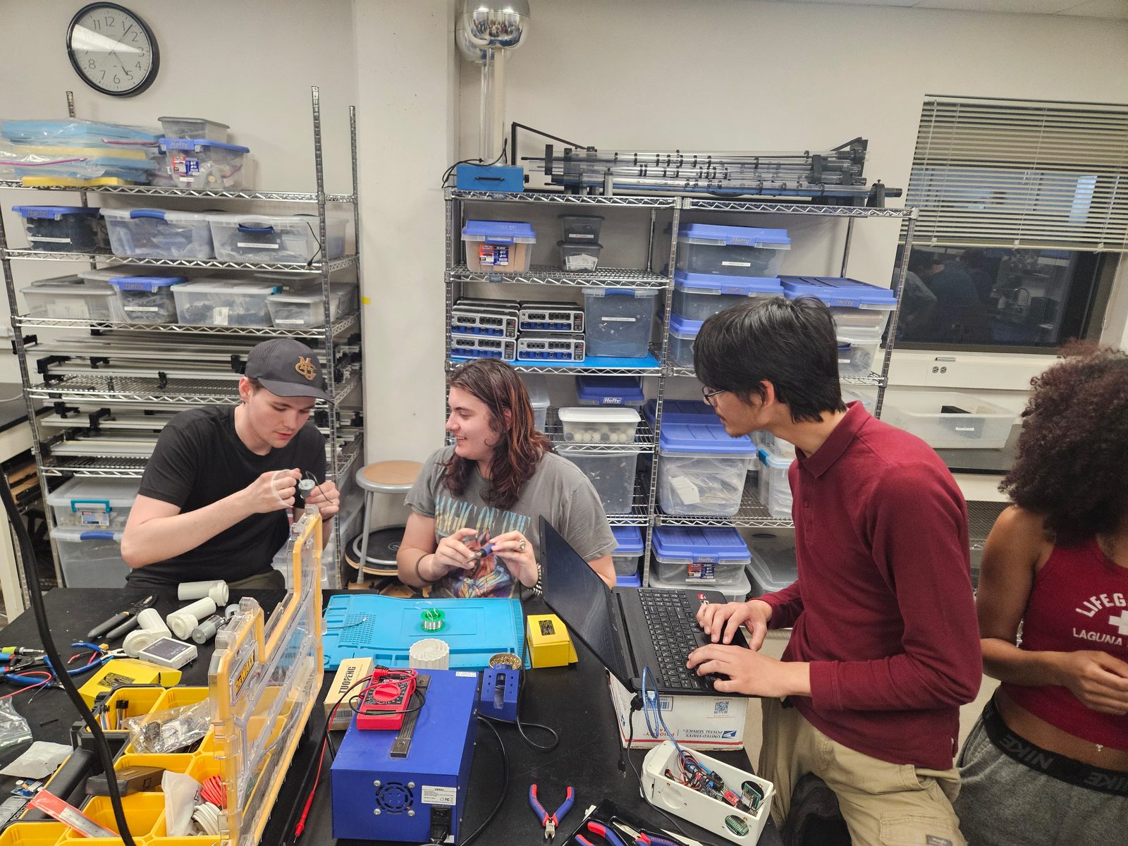 Four students work at a lab bench with a soldering station, multimeter and parts bins while one types on a laptop